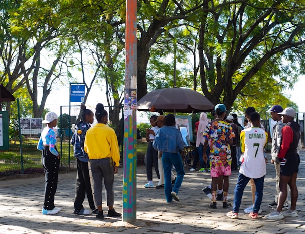 A group of people gathers in a park under leafy trees. They are interacting near a kiosk or stand surrounded by greenery and paved ground. Some individuals are wearing colorful clothing, and one person is wearing a number 7 sports jersey. There's a mixture of standing and walking activities happening in the scene.